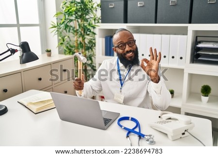 African american man working at medical clinic holding hammer doing ok sign with fingers, smiling friendly gesturing excellent symbol 