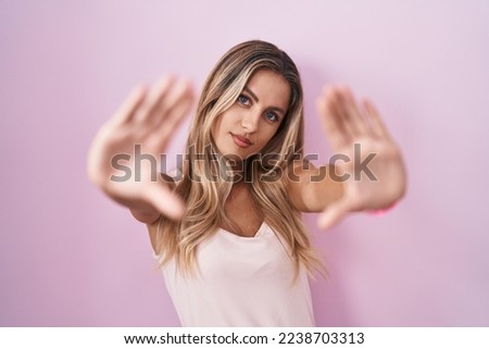 Young blonde woman standing over pink background doing frame using hands palms and fingers, camera perspective 