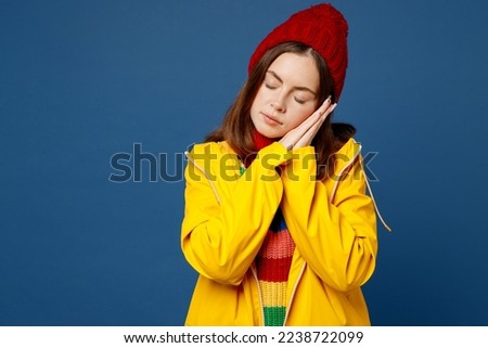 Young woman wear sweater red hat yellow waterproof raincoat outerwear hold folded hands under cheek close eyes isolated on plain dark royal navy blue background Outdoor wet fall weather season concept