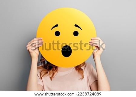 wow shocked smiley. Girl holds a yellow smiley with surprised face emoticon isolated on a grey studio background. Cartoon face.