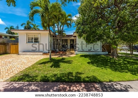 Corner house facade surrounded by trees and palm trees with parking spaces, short grass, tile roof, blue sky