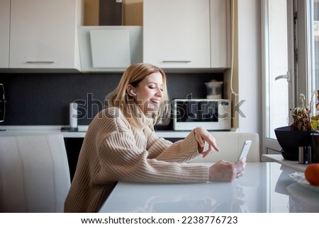 beautiful young woman in a cozy sweater sits in the kitchen and watches updates on social networks. Girl at home with phone in hand