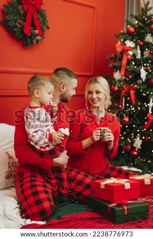 Happy Christmas Family Father Mother and Baby Son sitting near Christmas tree and having fun. Red background and red and green gift boxes. Woman holding golden cup with marshmallow 