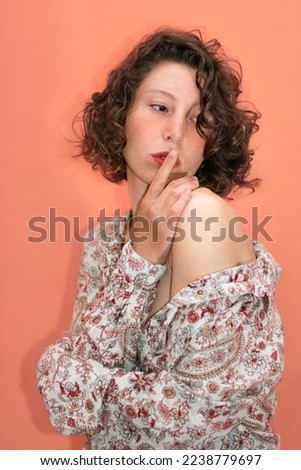studio shot of a beautiful young woman with curlers and short brown hair posing confidently. The background is pink-orange