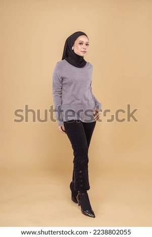 Full length portrait of a beautiful Muslim female model wearing grey and black attire with hijab isolated over beige  studio background. Fashion, beauty, lifestyle concept.