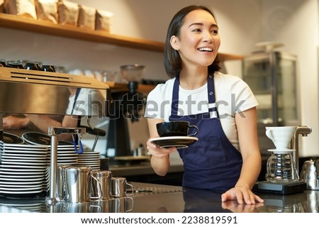Portrait of beautiful asian girl, student working part-time in cafe, holding cup of coffee, made order, looking for client, wearing uniform. Royalty-Free Stock Photo #2238819591