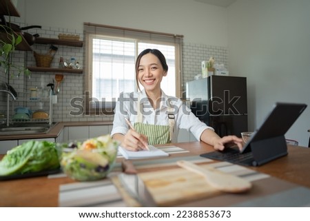 Young beautiful woman using a laptop computer searching and learning for cooking healthy food from fresh vegetables and fruits in the kitchen room.