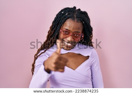 African woman with braided hair standing over pink background doing happy thumbs up gesture with hand. approving expression looking at the camera showing success. 
