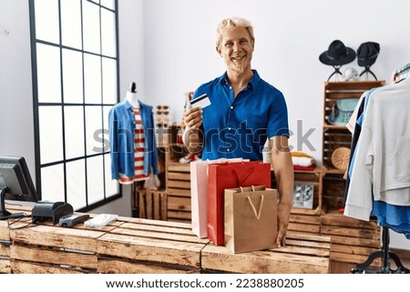 Young blond man holding credit card working as manager at retail boutique looking positive and happy standing and smiling with a confident smile showing teeth 