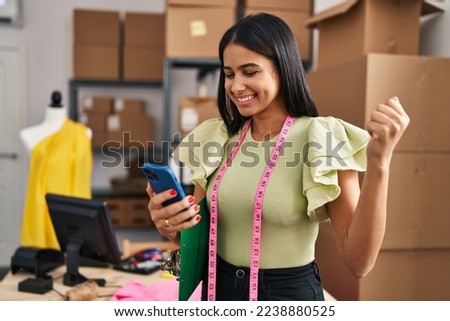 Young latin woman dressmaker with winner expression using smartphone at designer studio Royalty-Free Stock Photo #2238880525