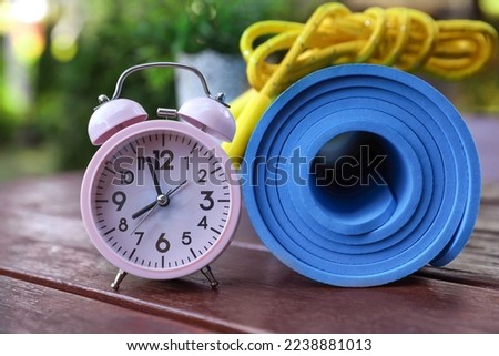 Alarm clock, fitness mat and skipping rope on wooden table outdoors, closeup. Morning exercise