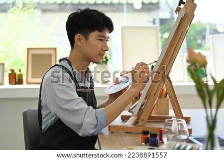 Concentred man painting picture on canvas with oil paints in bright art studio. Leisure activity and art concept