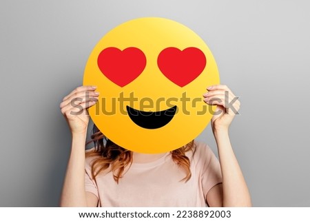 Emoticon in love. Girl holds a yellow smiley with romantic love face emoticon isolated on a grey background. Valentine's day concept
