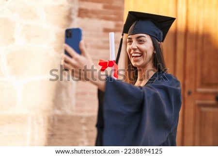 Young hispanic woman wearing graduated uniform holding diploma make selfie by the smartphone at university