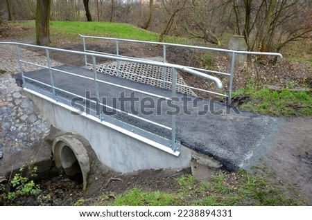 newly renovated bridge in the park over a small stream. concrete structures with a pipe passing through the flow lines. asphalt surface, metal sheet zinc drip tray. pipe railings, lawn, garden, cycle