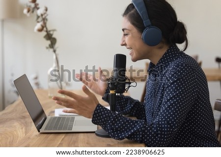 Happy pretty Indian blogger girl in blue wireless headphones speaking at laptop and professional microphone, talking on making video call, recording podcast, gesturing, smiling, laughing