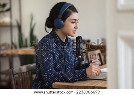 Calm pretty Indian radio host in wireless headphones broadcasting on air, speaking at studio big microphone, using laptop. Blogger, influencer, teacher, coach giving workshop, online conference
