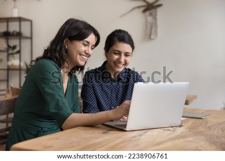Two cheerful diverse office workers women cooperating on project, sitting at work desk with laptop, looking at screen, smiling, laughing. Female mentor teaching Indian student, pointing at monitor Royalty-Free Stock Photo #2238906761