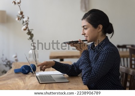 Positive young freelance Indian business woman using app on gadgets for online communication, recording voice message on smartphone, pointing at laptop computer monitor, working at desk in home office