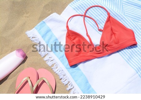 Striped towel with bikini top, bottle of sunblock and flip flops on sandy beach, flat lay