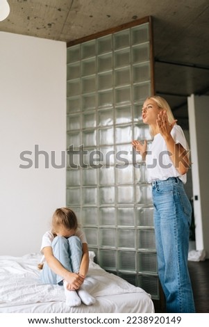 Furious blonde mom scolding, raising voice, yelling, gesturing with hands to crying little girl covering face with palm sitting on bed at home. Concept of crisis in family, domestic violence, abuse.
