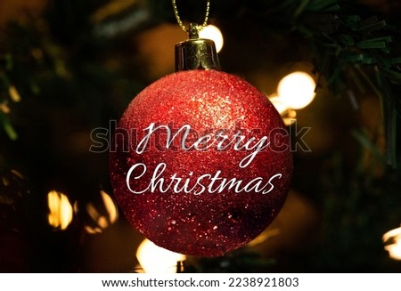 Red Shiny Christmas Ball Decoration with Merry Christmas Text in Christmas Lights. Closeup of Red Christmas Ornament with Text Royalty-Free Stock Photo #2238921803