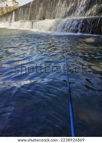 fishing with a blue rod from the edge of the shady Gumelem river during the day, the waterfall from the dam stretches