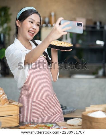 Young woman wears apron taking selfie photo with homemade pie in kitchen. Portrait of beautiful Asian female baking dessert and having fun taking photo by smartphone for online social. Home cookery.