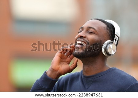Happy black man wearing wireless headphones listening to music laughing in the street