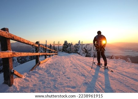 A skier is resting on a ski slope with beautiful view to the snowy mountains and stay to admire the surrounding landscape and beautiful sunset.Photographed in natural light against sunset light.