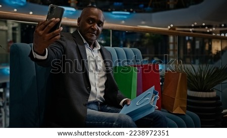 Middle-aged male African man buyer client customer blogger influencer vlogger with shopping bags sitting in mall video call make photography with purchase smartphone photographing phone photo picture