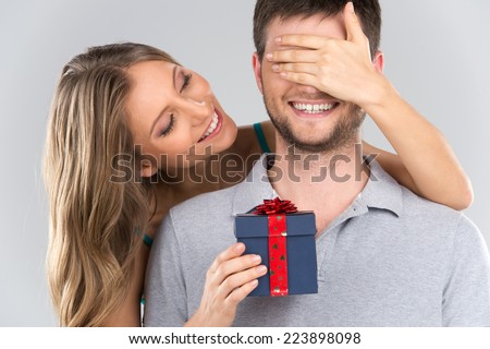 romantic woman covering her boyfriend's eyes. girl standing behind man with gift isolated on grey background