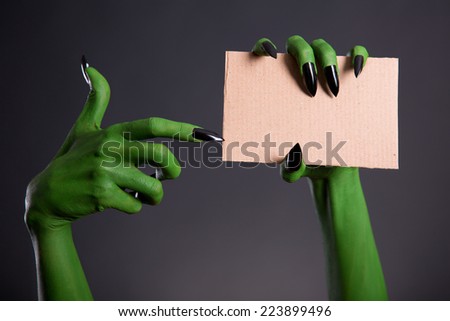Green monster hand with black nails pointing on blank piece of cardboard, Halloween theme  