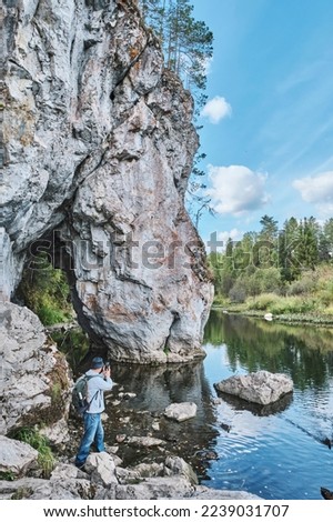 Senior male hiker with backpack taking pictures on mobile phone on bank of Serga River near rock Holey stone. Rear view. Summer nature landscape. Nature park Deer streams. Sverdlovsk region, Russia