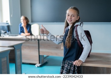 Caucasian schoolgirl with a backpack and a female teacher in the classroom on showing thumbs up.