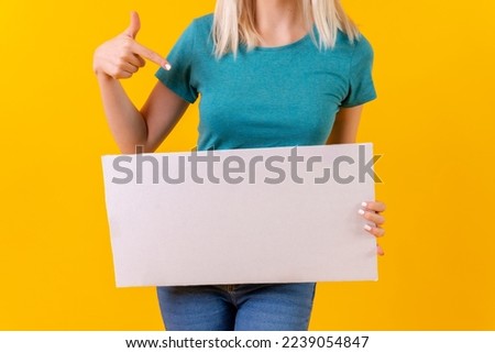 Pointing at white empty advertisement poster, blonde caucasian girl on yellow studio background
