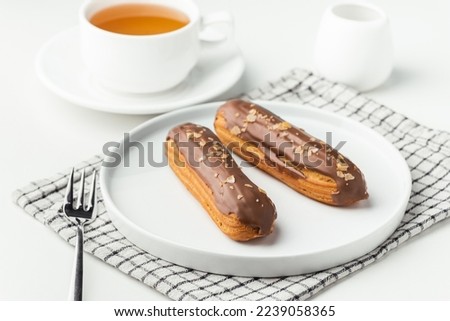 Eclairs with custard and chocolate icing on a white plate. Eclairs with cream filling covered with melted milk chocolate. A cup of tea with traditional French pastry in the morning. French breakfast Royalty-Free Stock Photo #2239058365