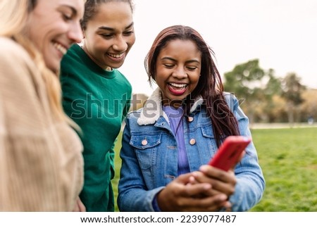Three multiracial female friends holding mobile phone outdoor. Millennial people watching social media content on smartphone device screen standing in city park Royalty-Free Stock Photo #2239077487