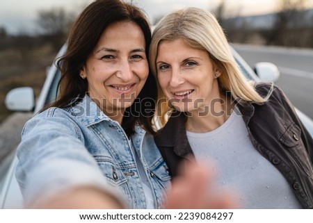 two women caucasian mature female friends taking selfie photos while standing outdoor in front to the car automobile on the road real people travel vacation concept copy space