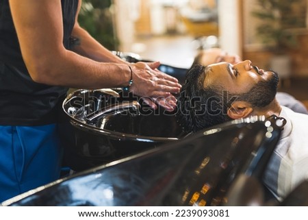 Indian male client having his hair washed by hairdresser next to blonde female customer in a professional hair salon. High quality photo