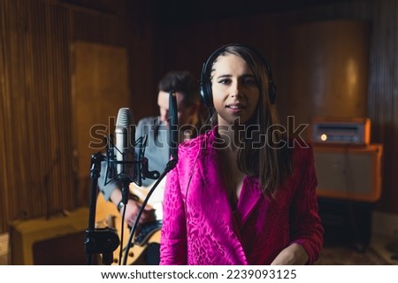 a soloist young woman with headphones standing close to a microphone guitarist in the background, recording studio. High quality photo