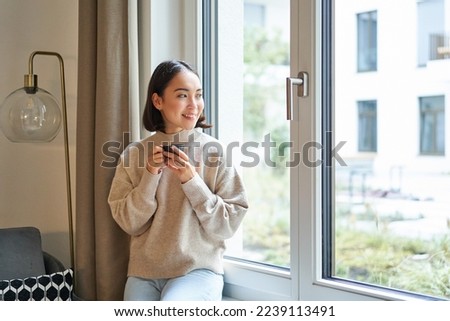 Beautiful young asian woman sitting near window and drinking her coffee, holding espresso cup and looking outside with relaxed, smiling face expression. Royalty-Free Stock Photo #2239113491