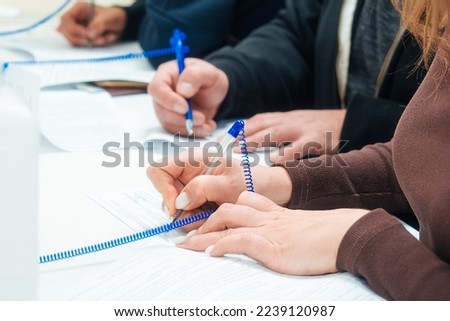 Woman fills out blank application form at table or signs contract. Hands of people with close-up fountain pen. Perspective view Several people simultaneously fill in data with pen on paper..