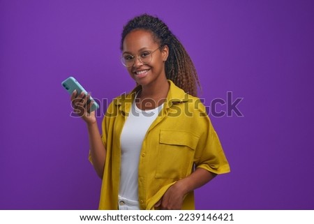 Young joyful pretty African American woman with phone smiling and looking at camera using applications for online dating or chatting with friends in instant messengers stands on lilac background Royalty-Free Stock Photo #2239146421