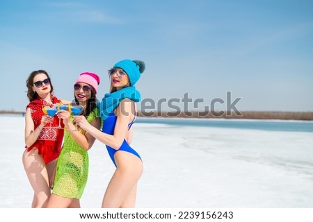 Three female friends in swimsuits drink blue alcoholic cocktails on a snowy beach. Hot girls posing in bikinis outdoors in winter.