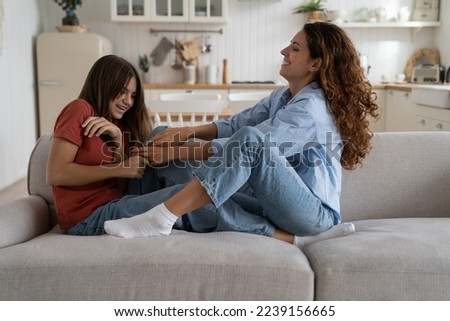 Smiling casual mom reaching out hands to teenage girl to tickle baby sits on sofa. Caucasian woman tickling laughing daughter for happy childhood concept. Love in the family. 