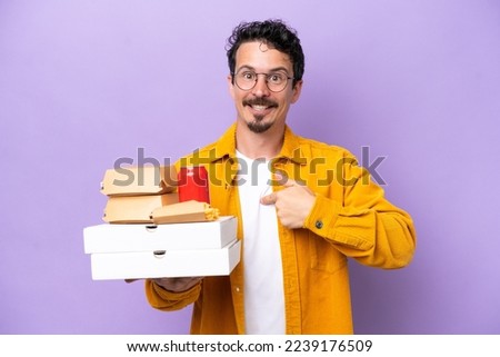 Young caucasian man holding fast food isolated on purple background with surprise facial expression