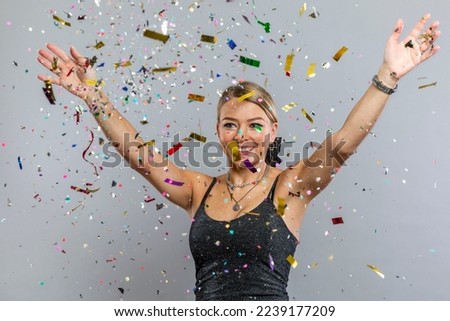 Young woman in confetti. Laughing blonde in a smart black dress rejoices in a surprise on a holiday. Happy New Year and Merry Christmas. Gray background.