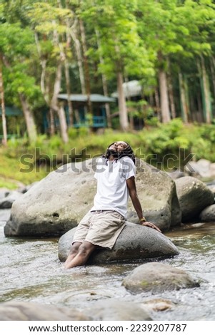 A young man with dreadlocks wearing a blank white shirt sitting while enjoying his surrounding near a river in the nature