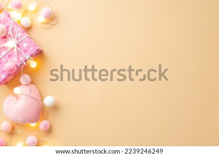 St Valentine's Day concept. Top view photo of giftbox soft heart shaped toy light bulb garland and fluffy pompons on isolated pastel beige background with blank space